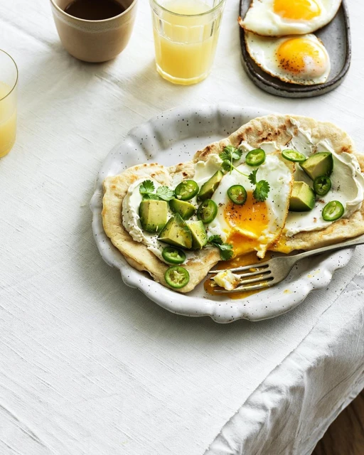 Overhead shot of two breakfast plates with sunny-side-up eggs, avocado, and sliced jalapeños on a white linen tablecloth.