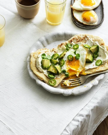 Overhead shot of two breakfast plates with sunny-side-up eggs, avocado, and sliced jalapeños on a white linen tablecloth.