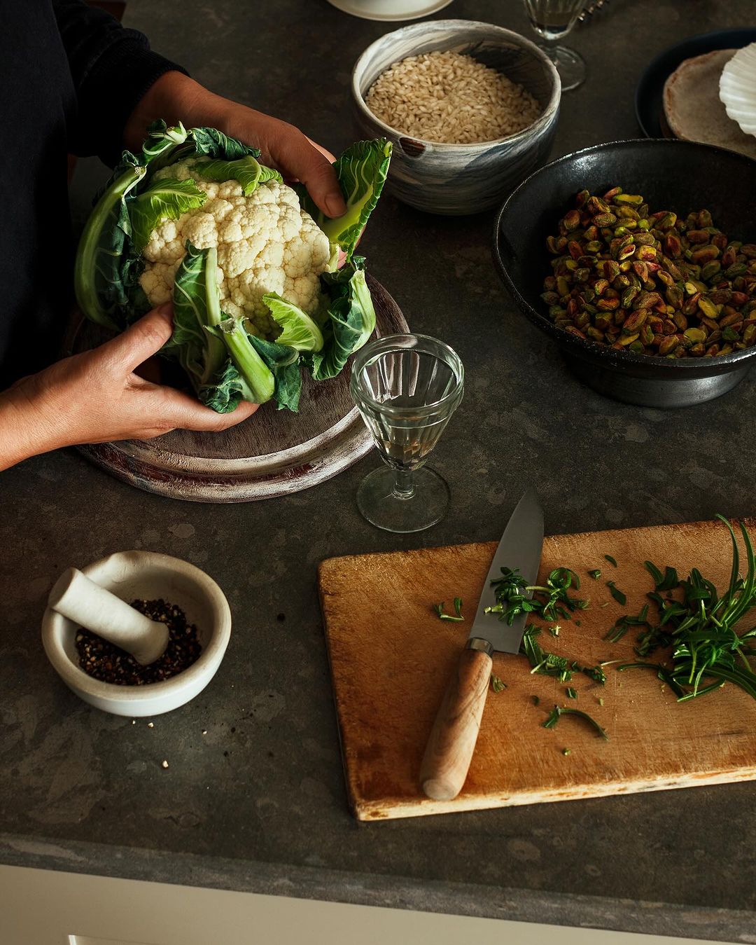 Person preparing cauliflower with herbs and spices on a wooden board in a kitchen, ingredients in bowls and natural light.