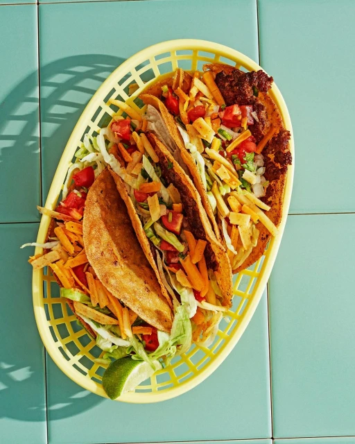 Overhead shot of three tacos with lime in yellow plastic basket on tile surface featuring tortilla shells, shredded lettuce, and toppings.