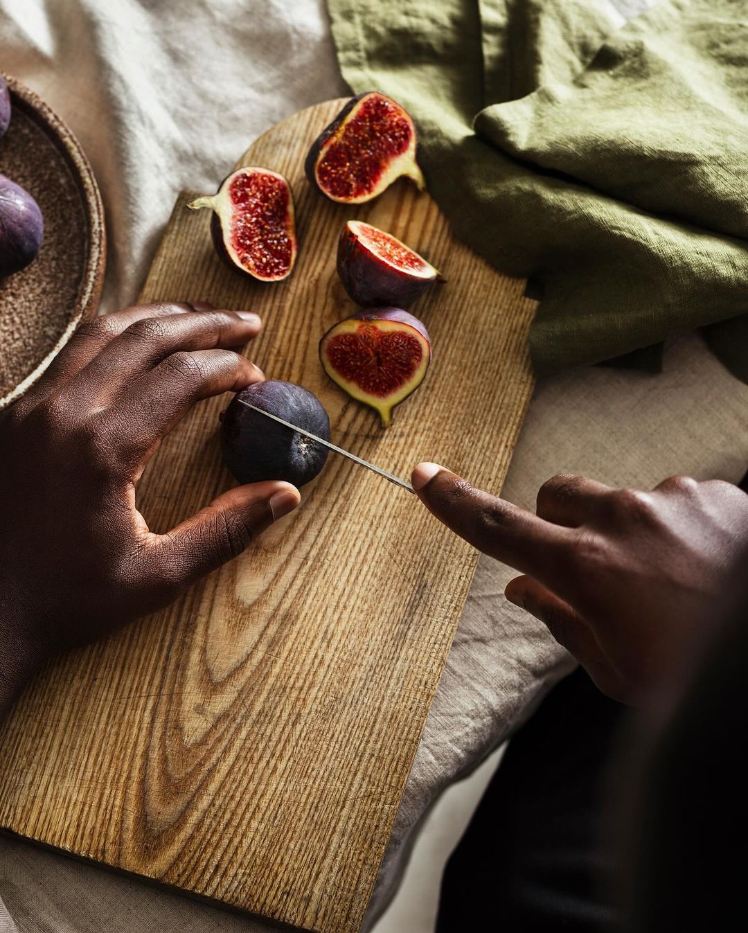 Overhead shot of figs being cut on a wooden board by a person with dark skin, with a gray linen cloth and green cloth in the background.