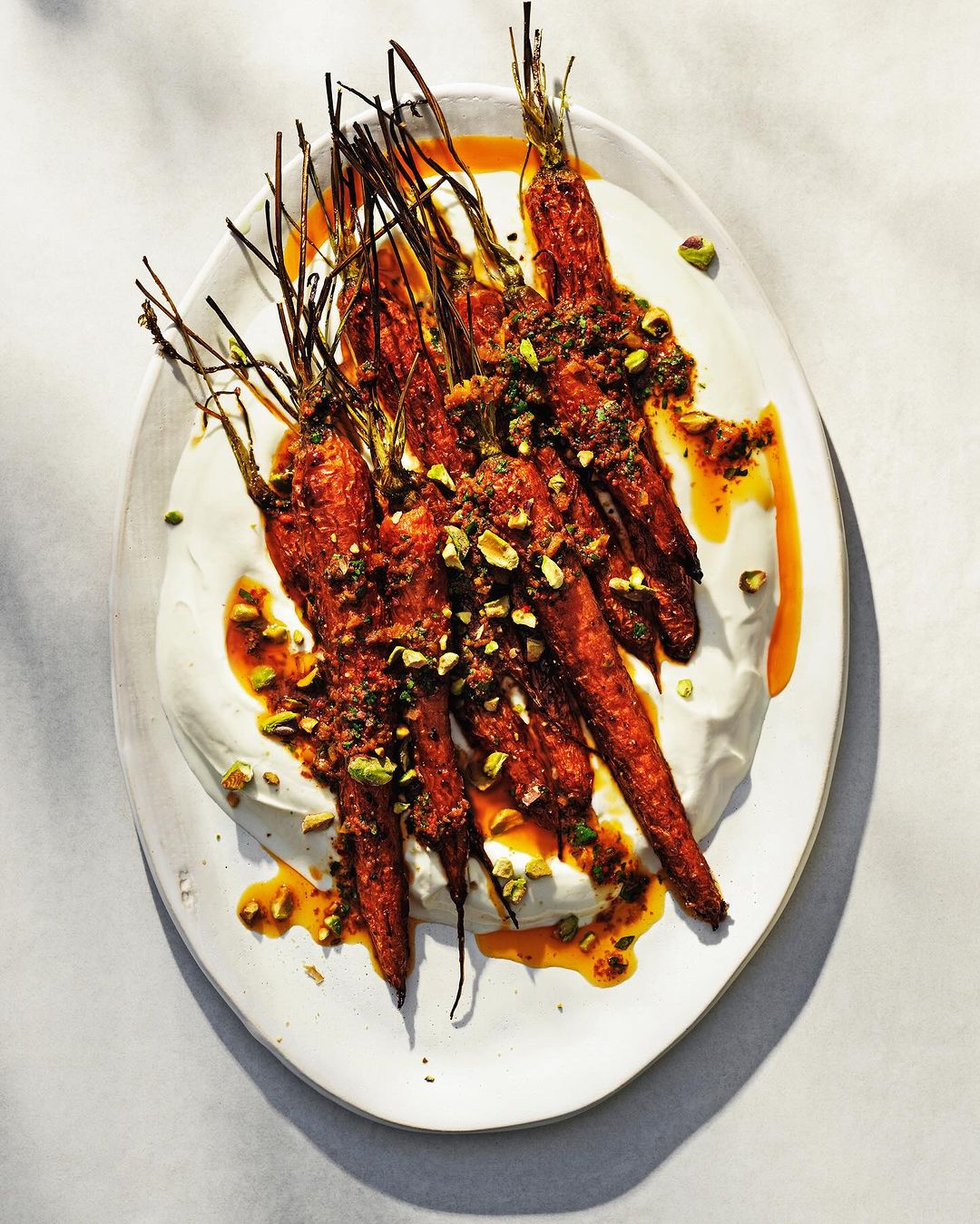 Overhead shot of roasted carrots topped with herbs and pistachios, served on a white plate with a white cream sauce and orange-brown glaze.