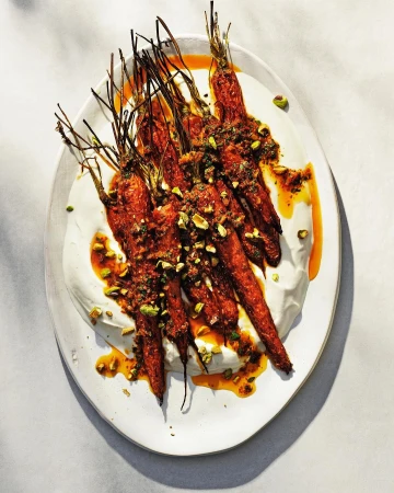 Overhead shot of roasted carrots topped with herbs and pistachios, served on a white plate with a white cream sauce and orange-brown glaze.