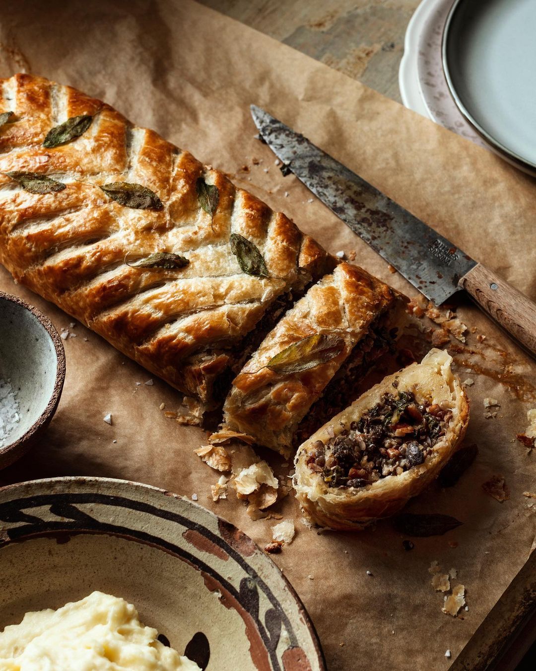 Overhead shot of a sliced mushroom Wellington with decorative sage leaves on parchment paper, plates, and a knife.
