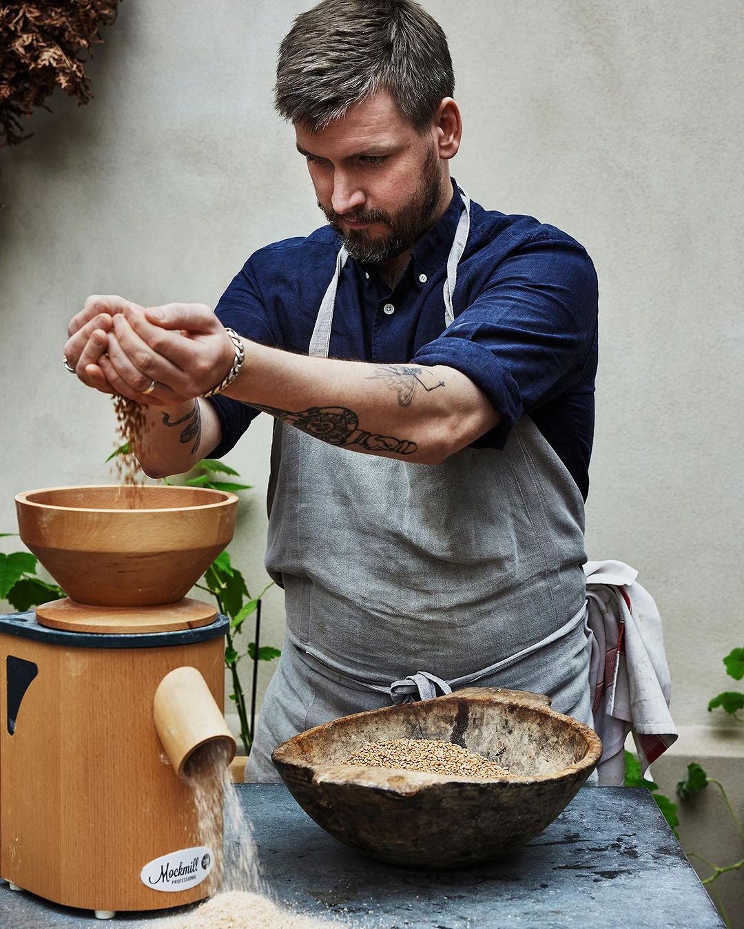 Adult man in a blue shirt and apron pours grain into a wooden mill in a natural light food setting with wooden bowls.