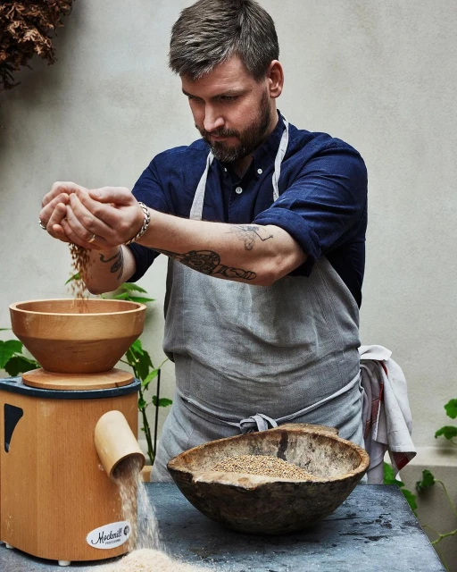 Adult man in a blue shirt and apron pours grain into a wooden mill in a natural light food setting with wooden bowls.