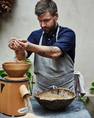 Adult man in a blue shirt and apron pours grain into a wooden mill in a natural light food setting with wooden bowls.