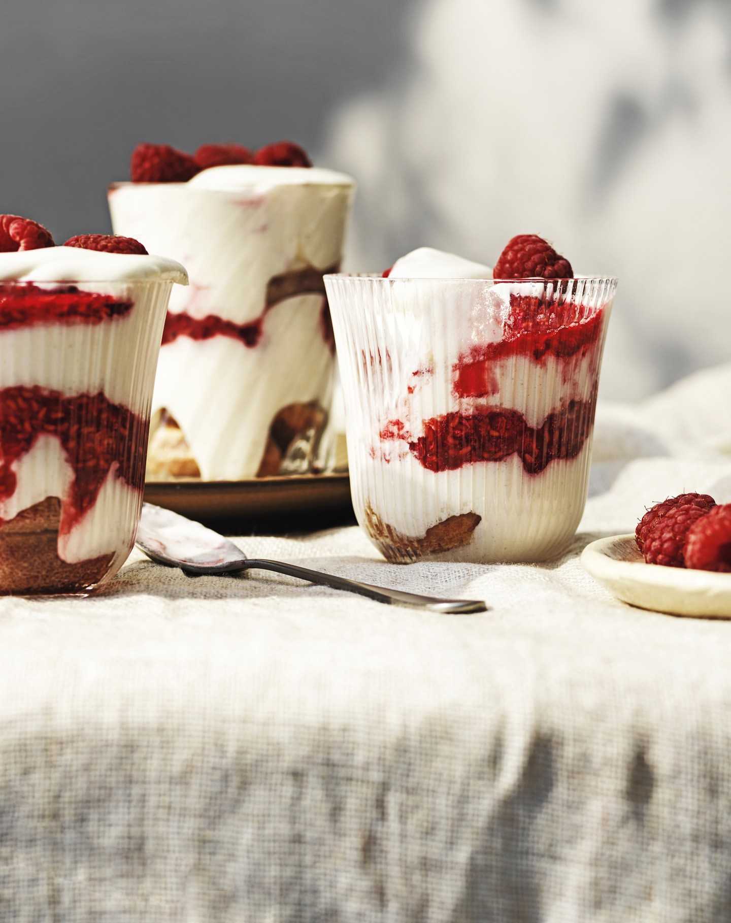 Three glasses filled with raspberry, cream, and cake layers on a beige linen tablecloth with a spoon and small plate of raspberries.