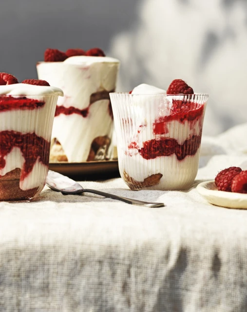 Three glasses filled with raspberry, cream, and cake layers on a beige linen tablecloth with a spoon and small plate of raspberries.