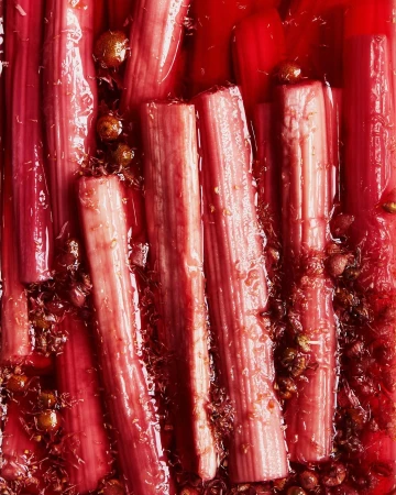 Close-up of several stalks of rhubarb poached in a vibrant red liquid, adorned with small, round golden-brown aromatics. Food photography.