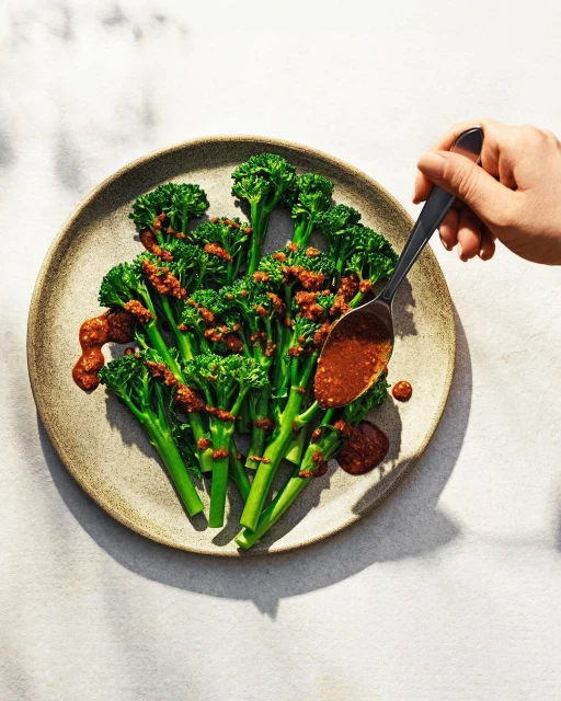 Overhead shot of cooked broccolini on a plate with red sauce, drizzled with a spoon, showcasing food presentation and culinary art.