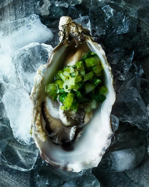 Overhead shot of a single oyster in its shell, filled with green relish and resting on a bed of crushed ice.