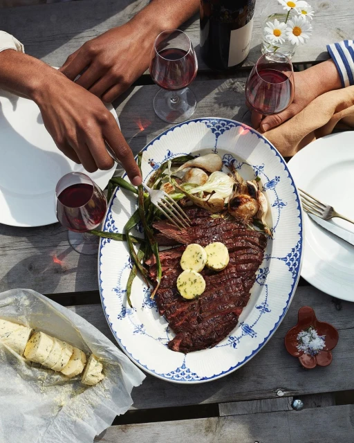 Overhead shot of steak with butter, onions, and greens on a plate surrounded by two people, glasses of wine, and wooden table.