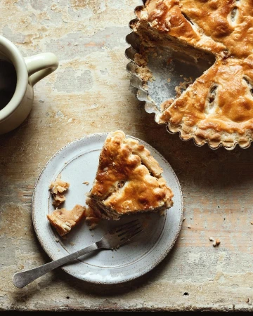 Overhead shot of a slice of crusty golden pie on a white plate with a fork, set on a rustic wooden table with coffee mug.