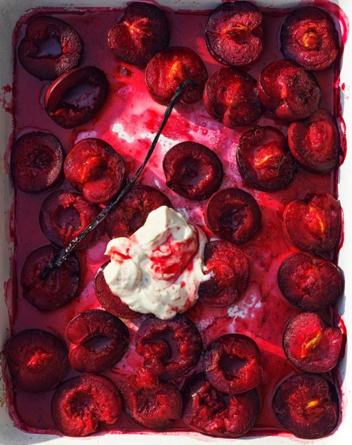 Overhead shot of halved plums in a white dish with cream and vanilla bean, arranged in rows, bathed in plum juice.