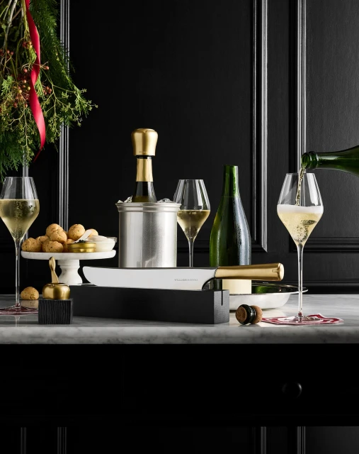 Champagne is being poured into a flute glass next to a wine cooler, cake stand, and knife set against a black paneled background.