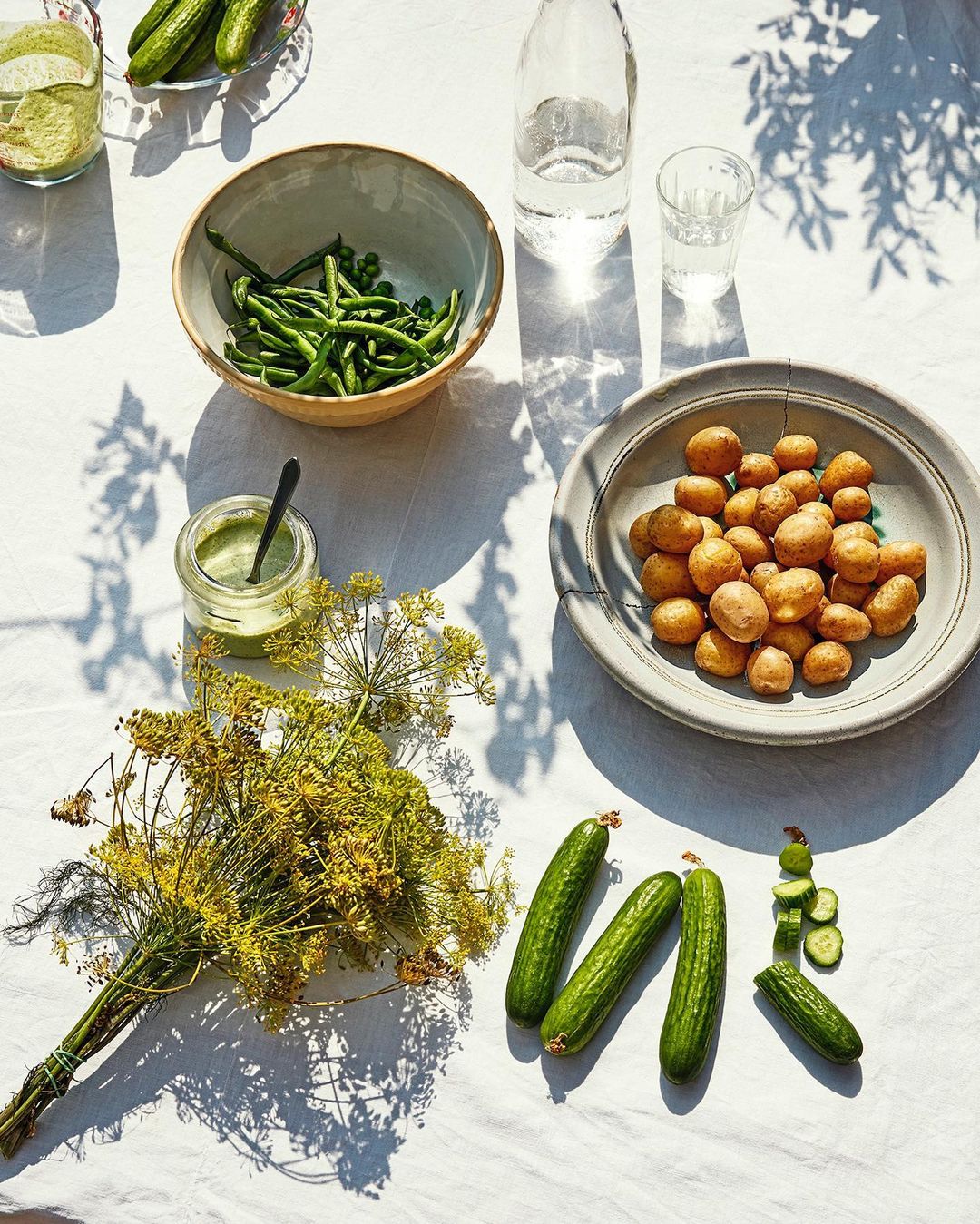 Overhead shot features fresh green beans, new potatoes, dill, cucumbers, and green sauce on a white tablecloth with natural sunlight and shadows.