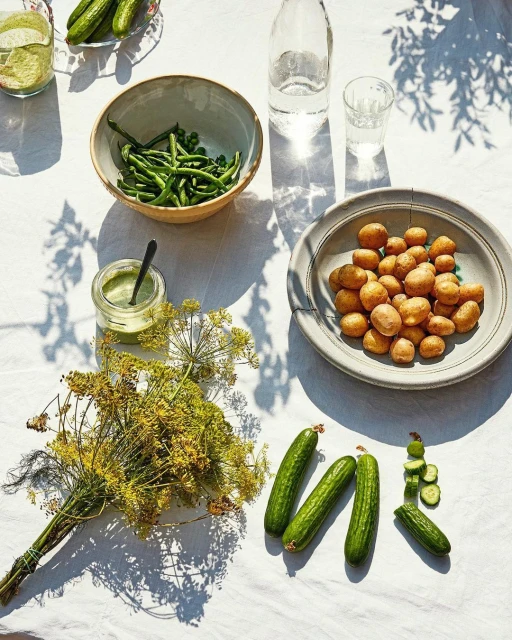 Overhead shot features fresh green beans, new potatoes, dill, cucumbers, and green sauce on a white tablecloth with natural sunlight and shadows.