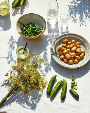Overhead shot features fresh green beans, new potatoes, dill, cucumbers, and green sauce on a white tablecloth with natural sunlight and shadows.