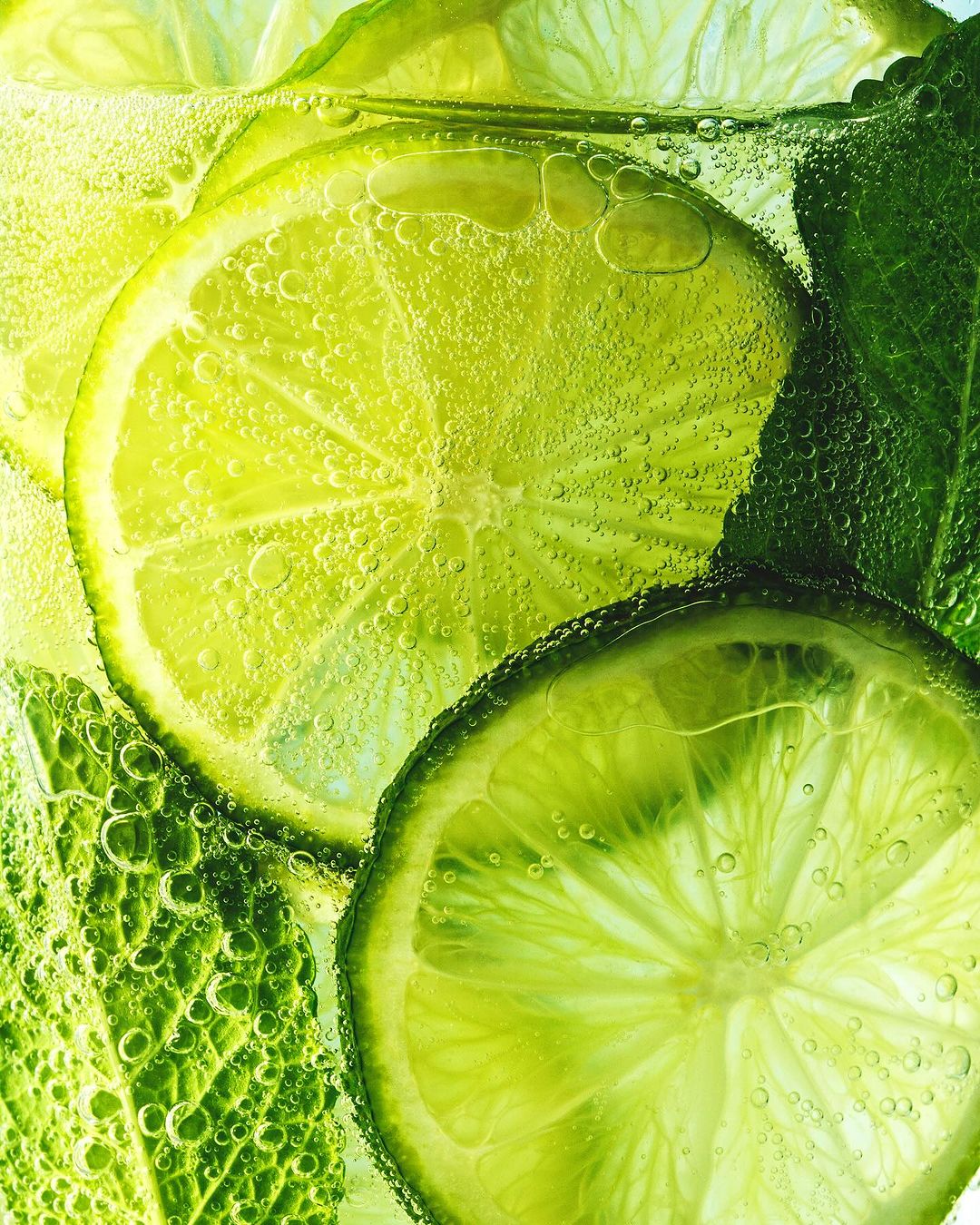 A close-up shot features lime slices and mint leaves suspended in a clear liquid, creating a refreshing and vibrant visual.