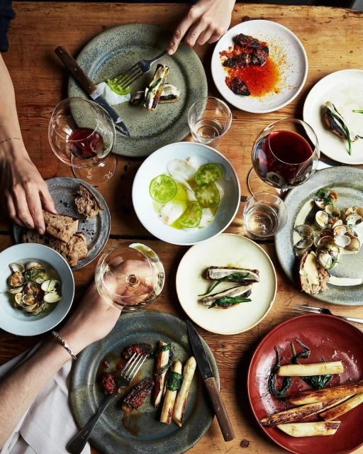 Overhead shot of a wooden table setting with plates of various food dishes, wine glasses, and hands reaching for the food, rustic presentation.