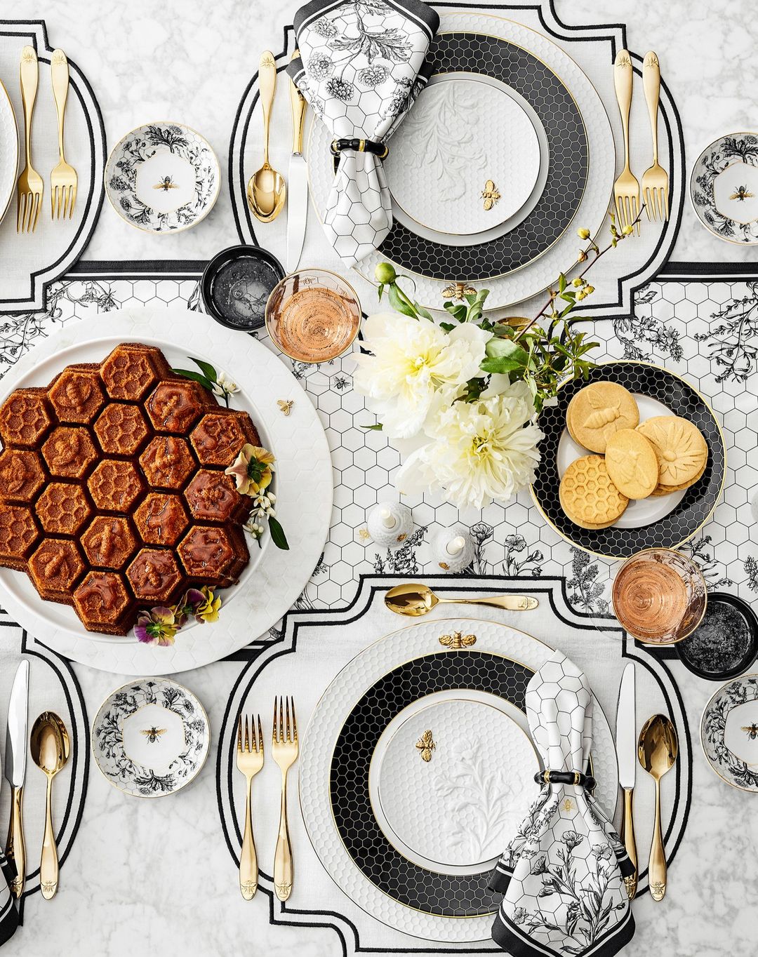 Overhead shot of a table setting with honeycomb cake, floral china plates with bee accents, gold flatware, and white floral centerpiece.