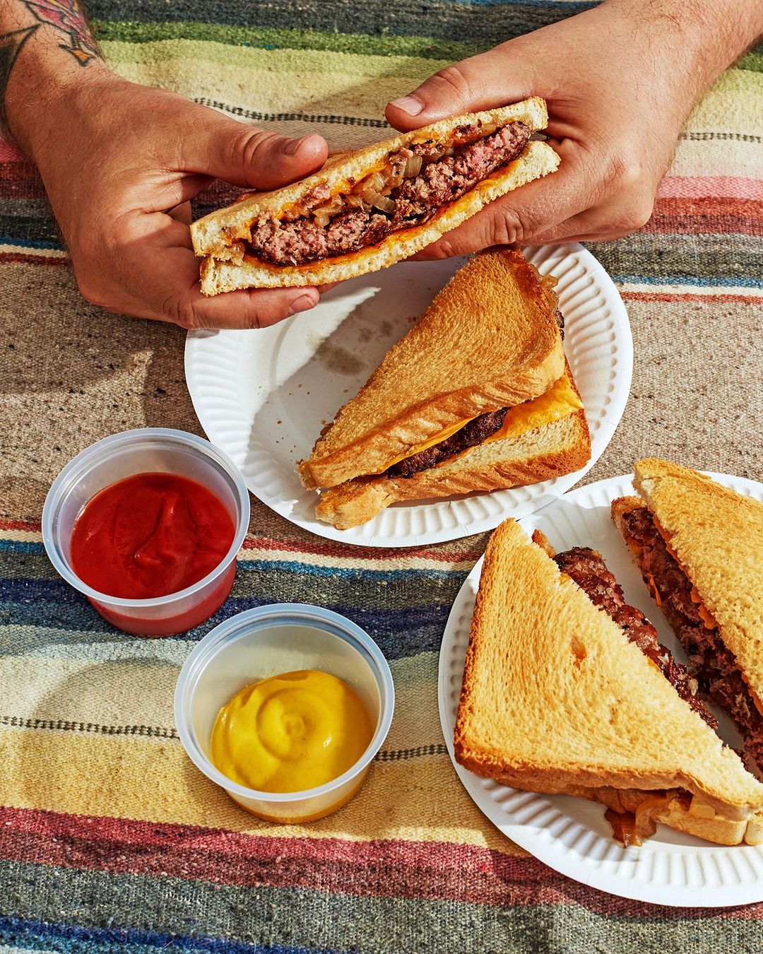 Overhead shot of two hands holding a sandwich over a plate with more sandwiches and condiments on a striped tablecloth.