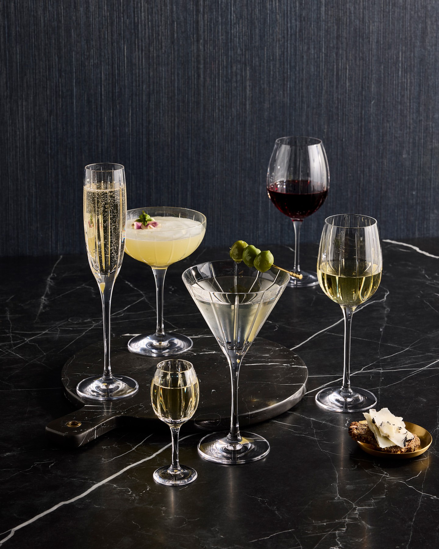 Several stemmed glasses with wine and cocktails are arranged on a black marble surface, some with olive garnish and cheese.