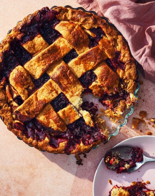 Overhead shot of a lattice-topped berry pie with a slice missing, served on a pink plate with a spoon, and a pink linen napkin.