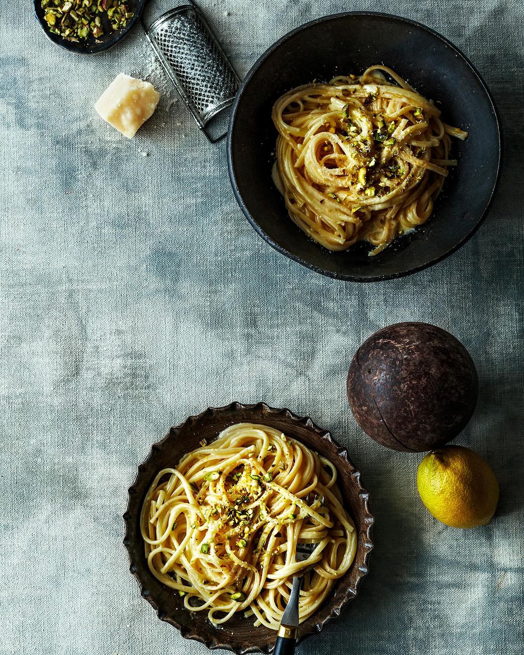 Overhead shot of two bowls of spaghetti with lemon, pistachio, and cheese, next to a lemon, cheese grater, and a small bowl of pistachios.