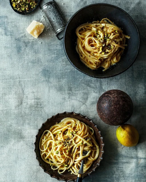 Overhead shot of two bowls of spaghetti with lemon, pistachio, and cheese, next to a lemon, cheese grater, and a small bowl of pistachios.