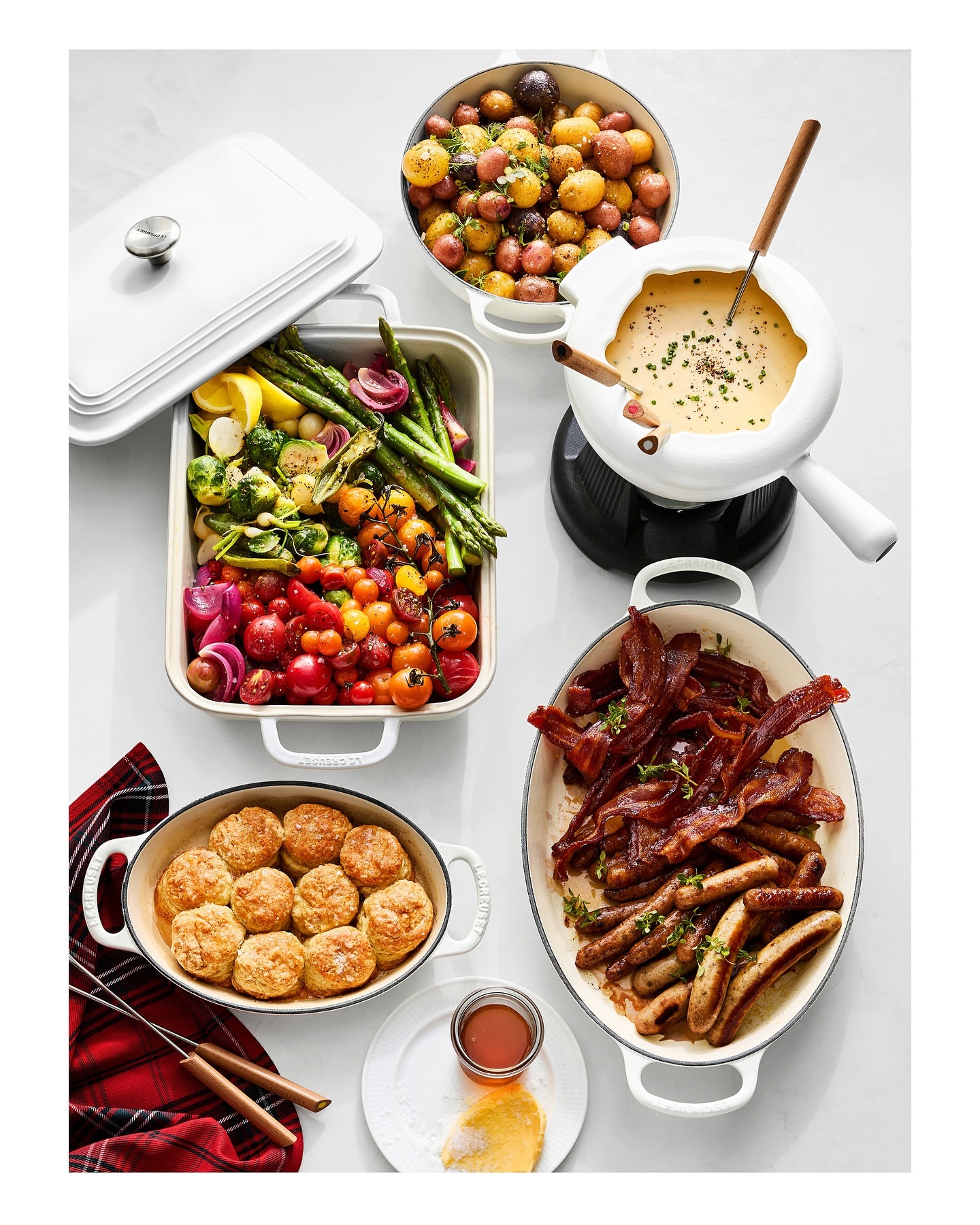 Overhead shot of a white table featuring platters of cooked vegetables, biscuits, bacon, sausages, potatoes, and cheese fondue.