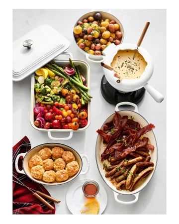 Overhead shot of a white table featuring platters of cooked vegetables, biscuits, bacon, sausages, potatoes, and cheese fondue.