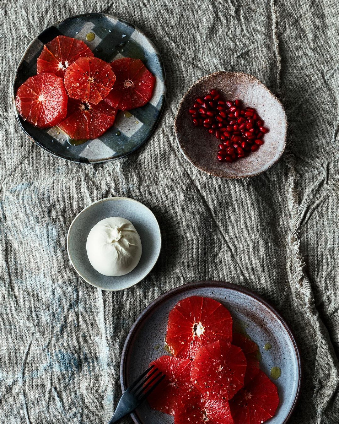 Overhead shot of a rustic linen surface displaying plates with vibrant blood orange slices, fresh burrata cheese, and pomegranate seeds.