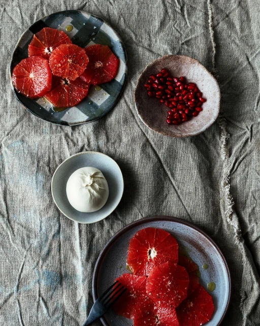 Overhead shot of a rustic linen surface displaying plates with vibrant blood orange slices, fresh burrata cheese, and pomegranate seeds.