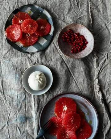 Overhead shot of a rustic linen surface displaying plates with vibrant blood orange slices, fresh burrata cheese, and pomegranate seeds.