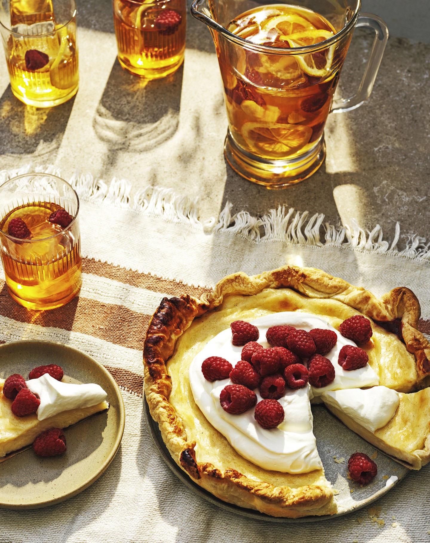 Overhead shot of a raspberry tart and a slice with a glass pitcher and glasses of tea with lemons and raspberries.