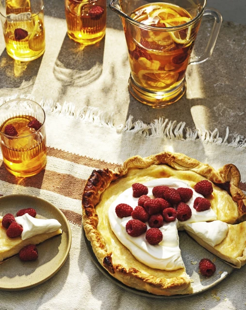 Overhead shot of a raspberry tart and a slice with a glass pitcher and glasses of tea with lemons and raspberries.