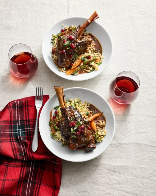 Overhead view of two plates featuring braised lamb shanks with couscous, carrots, and pomegranate seeds, alongside two glasses of red wine.