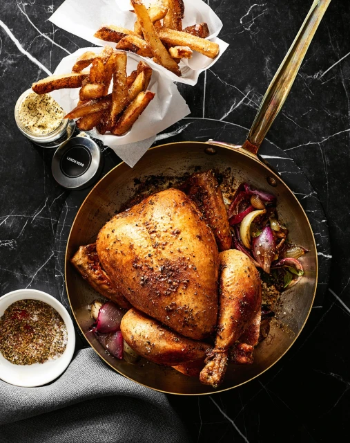 Overhead shot of a roasted chicken in a skillet with fries, onions, and condiments on a dark marble surface for food photography.