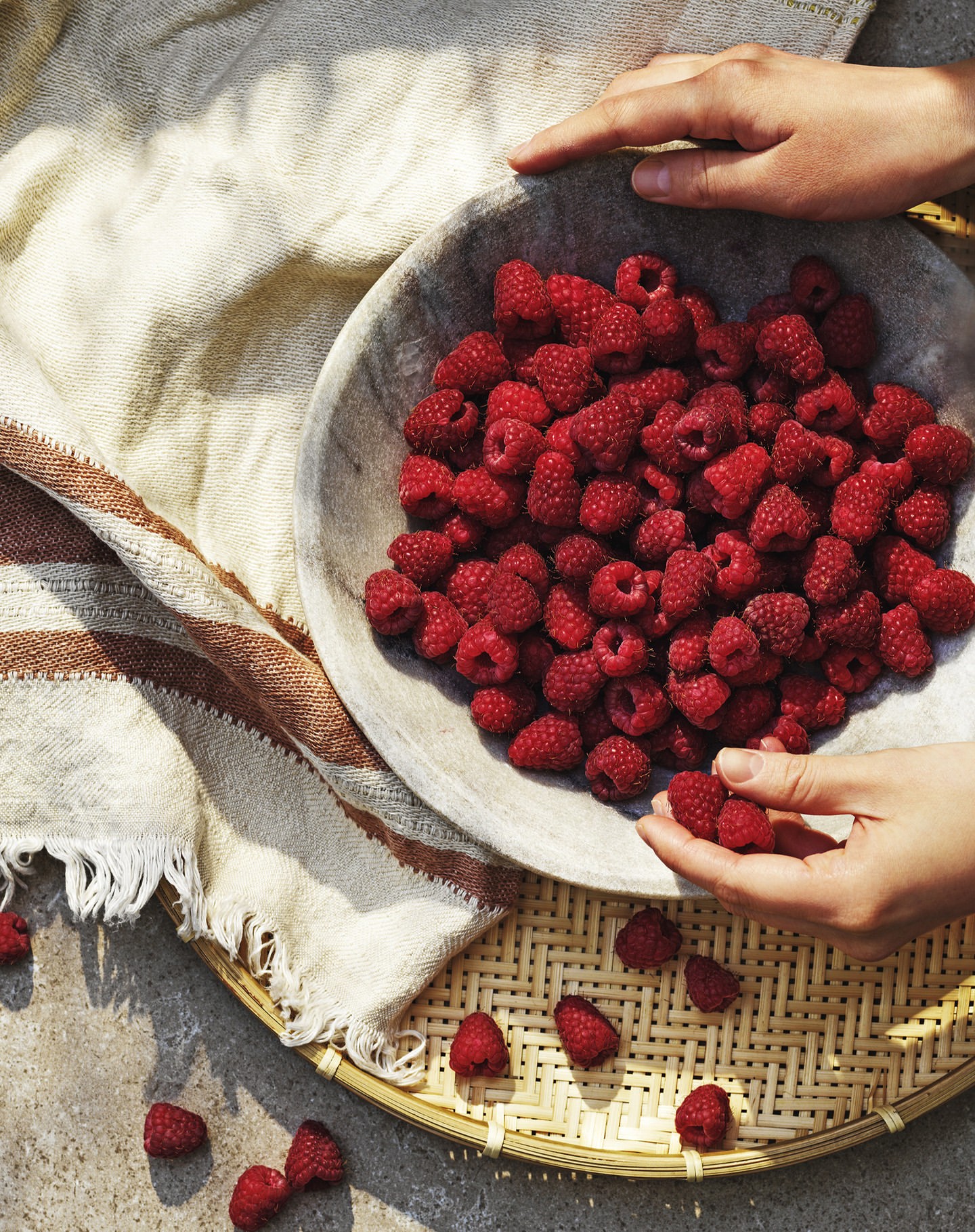 Overhead shot of hands arranging fresh raspberries in a rustic bowl, set on a woven tray with a striped linen cloth, and concrete surface.