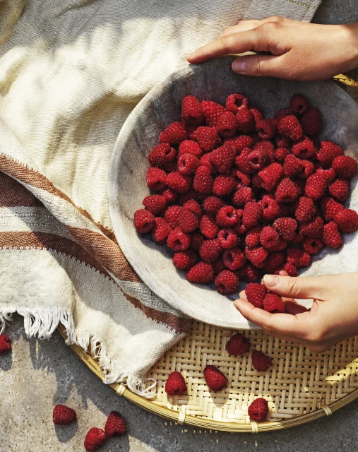 Overhead shot of hands arranging fresh raspberries in a rustic bowl, set on a woven tray with a striped linen cloth, and concrete surface.