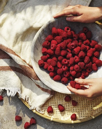 Overhead shot of hands arranging fresh raspberries in a rustic bowl, set on a woven tray with a striped linen cloth, and concrete surface.