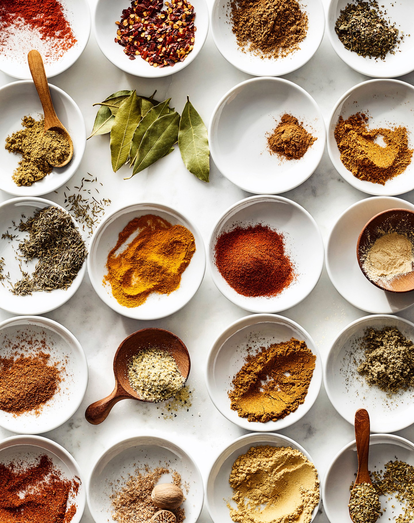 Overhead shot of spices and herbs arranged in white bowls on a white surface, creating a colorful and culinary-themed flat lay composition.