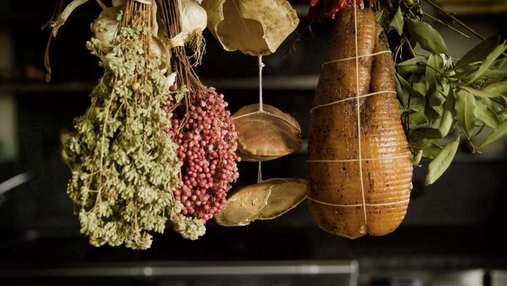 Hanging herbs and cured meats. A culinary still life with bay leaves, garlic, and strings of peppercorns in rustic, dark, indoor setting.