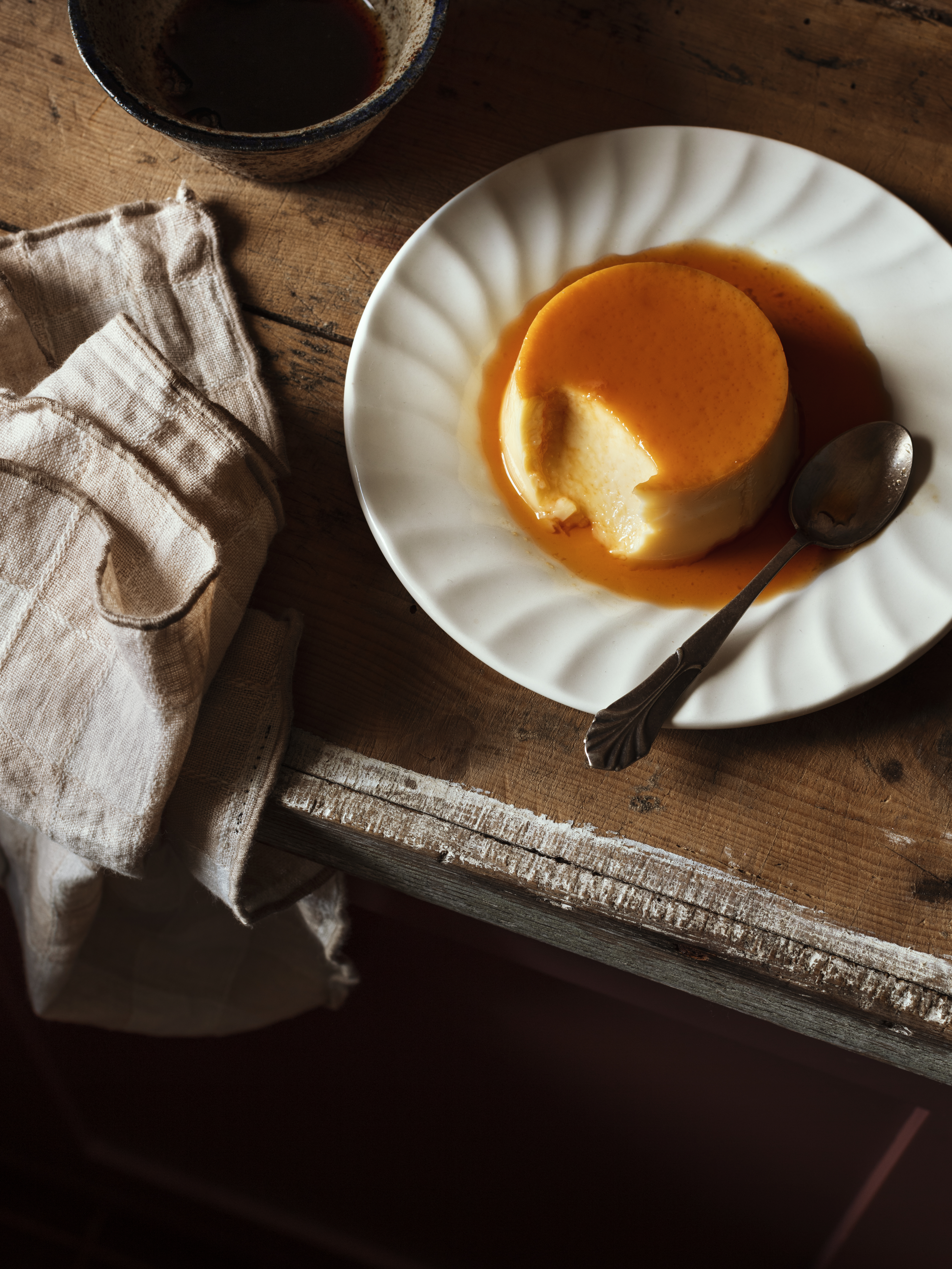 A custard flan dessert with a bite taken out, served on a white ridged plate with a silver spoon on a rustic wooden table.