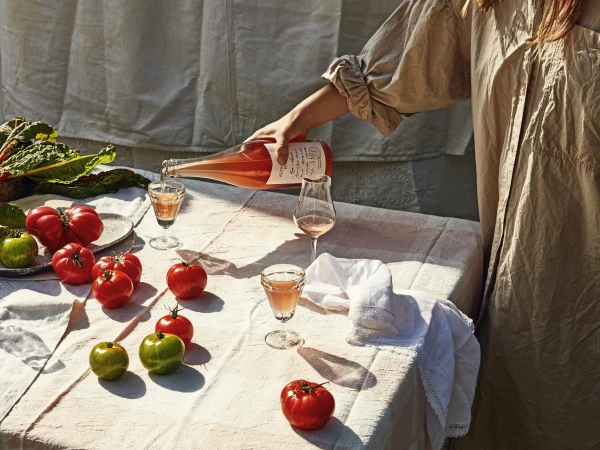 A person in a beige linen dress pours pink rosé wine from a bottle into a small glass on a table set with fresh heirloom tomatoes.