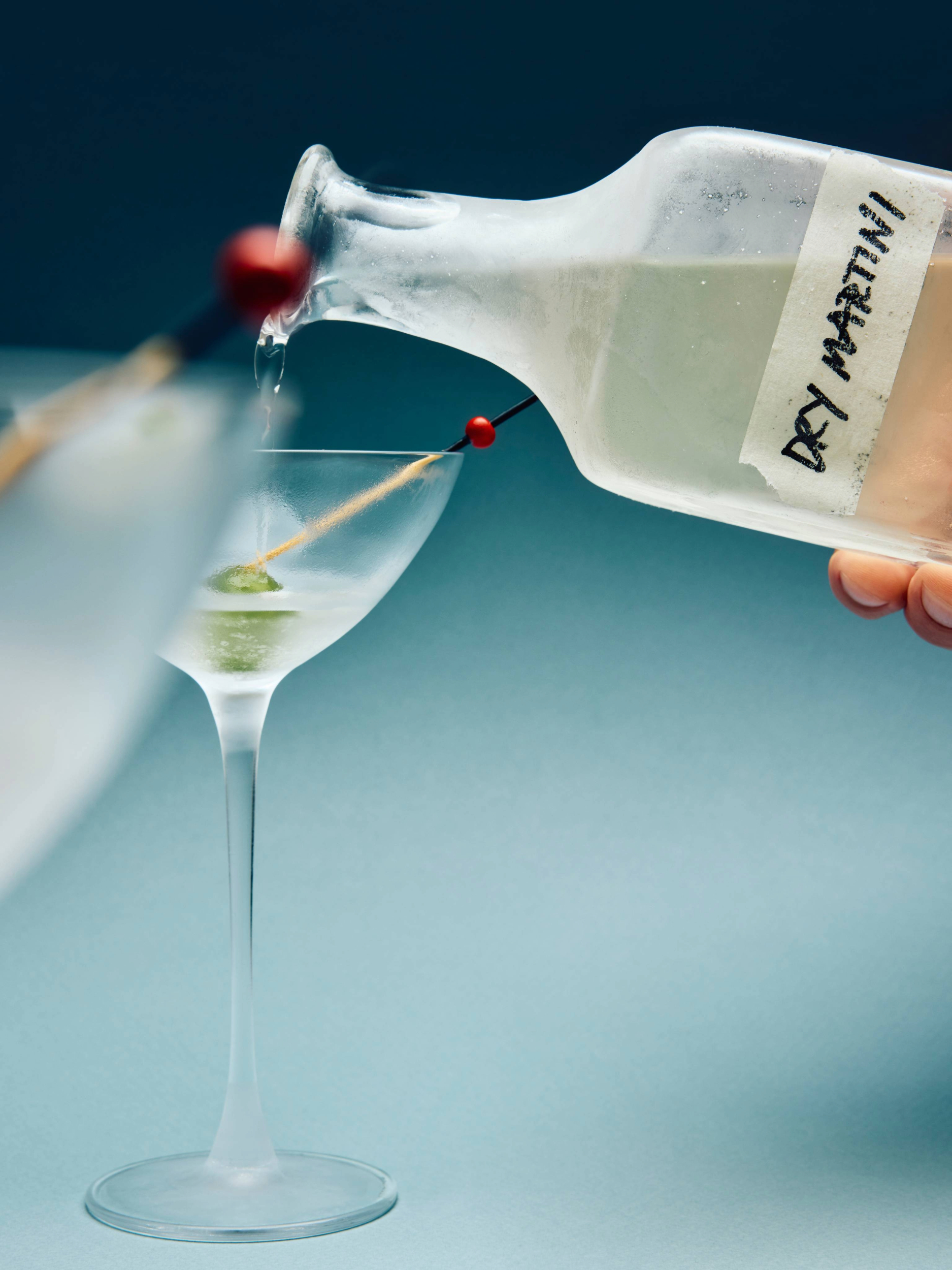 A hand pouring a dry martini from a frosted glass bottle into a chilled cocktail glass featuring a green olive on a pick.