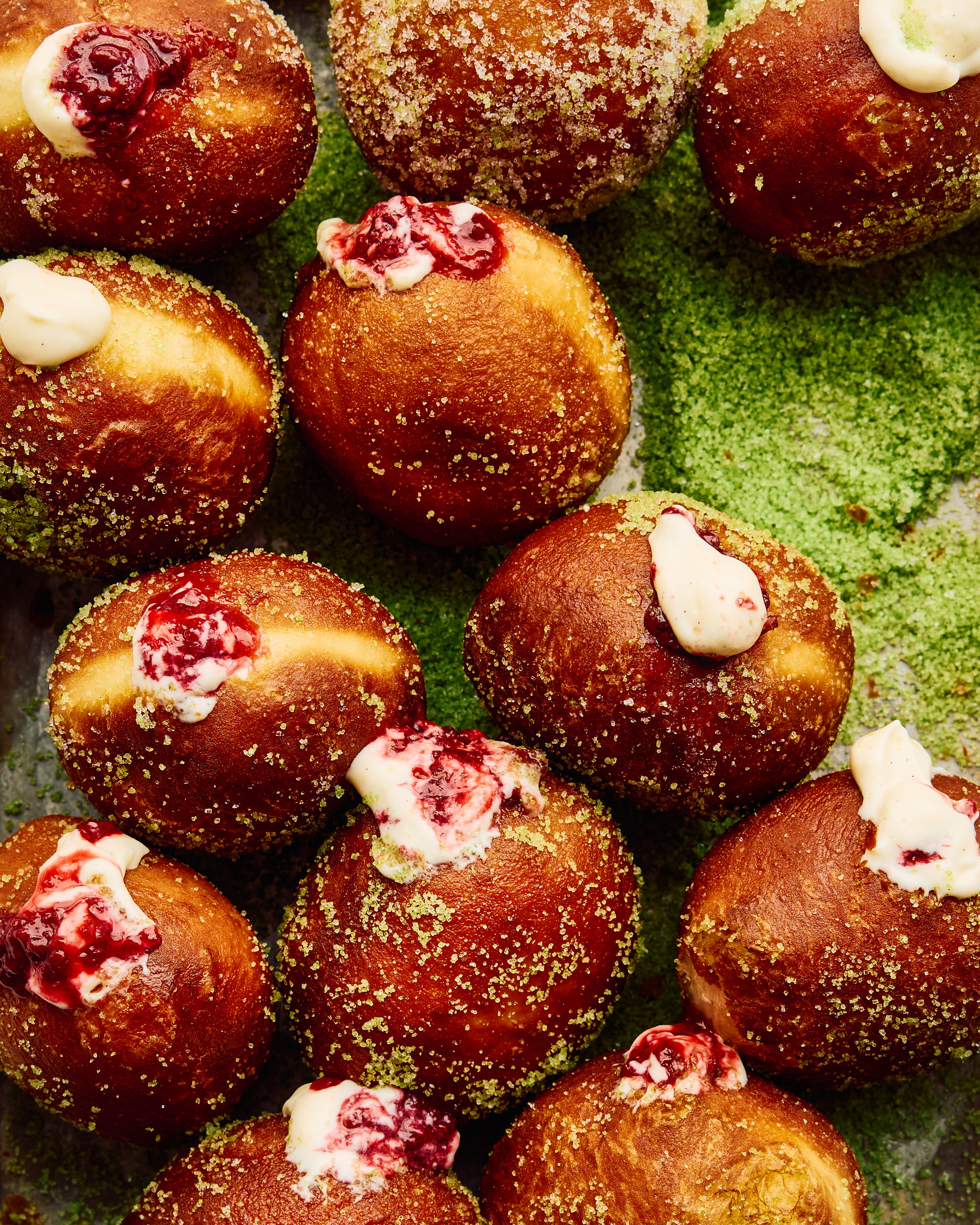 Top-down view of several round fried donuts filled with cream and berry jam, dusted with green matcha powder on a surface.