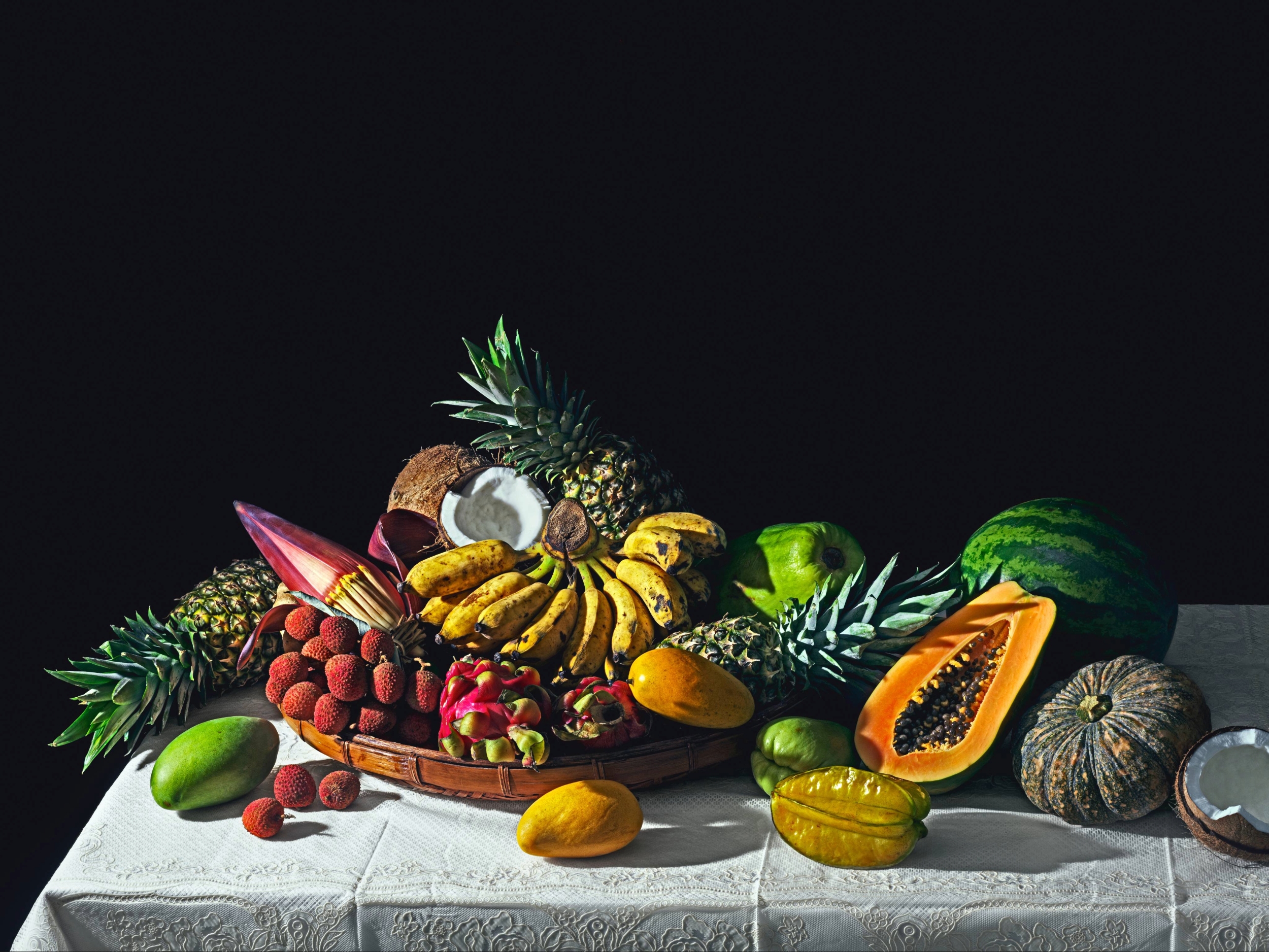 A still life composition of various tropical fruits including pineapple, mango, papaya, lychee, and coconut on a white tablecloth.